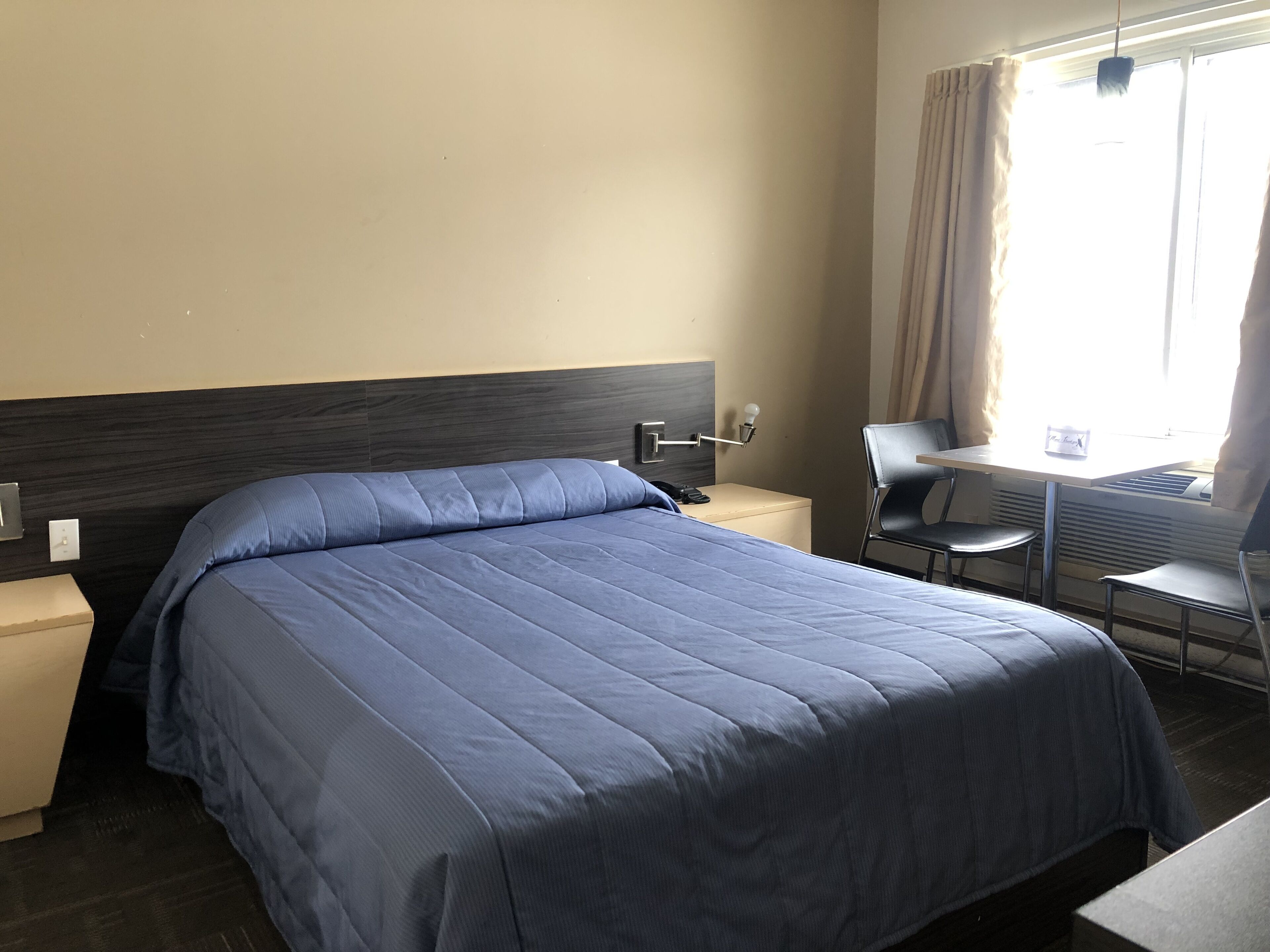 A hotel room with a bed, bedside table, lamp, chair, and a small table with a lamp.