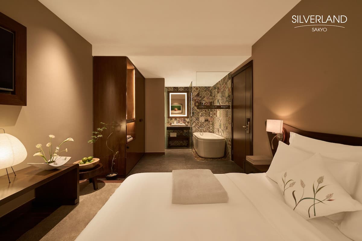 A modern hotel room with a bed, bedside table, lamp, and a view into the bathroom.