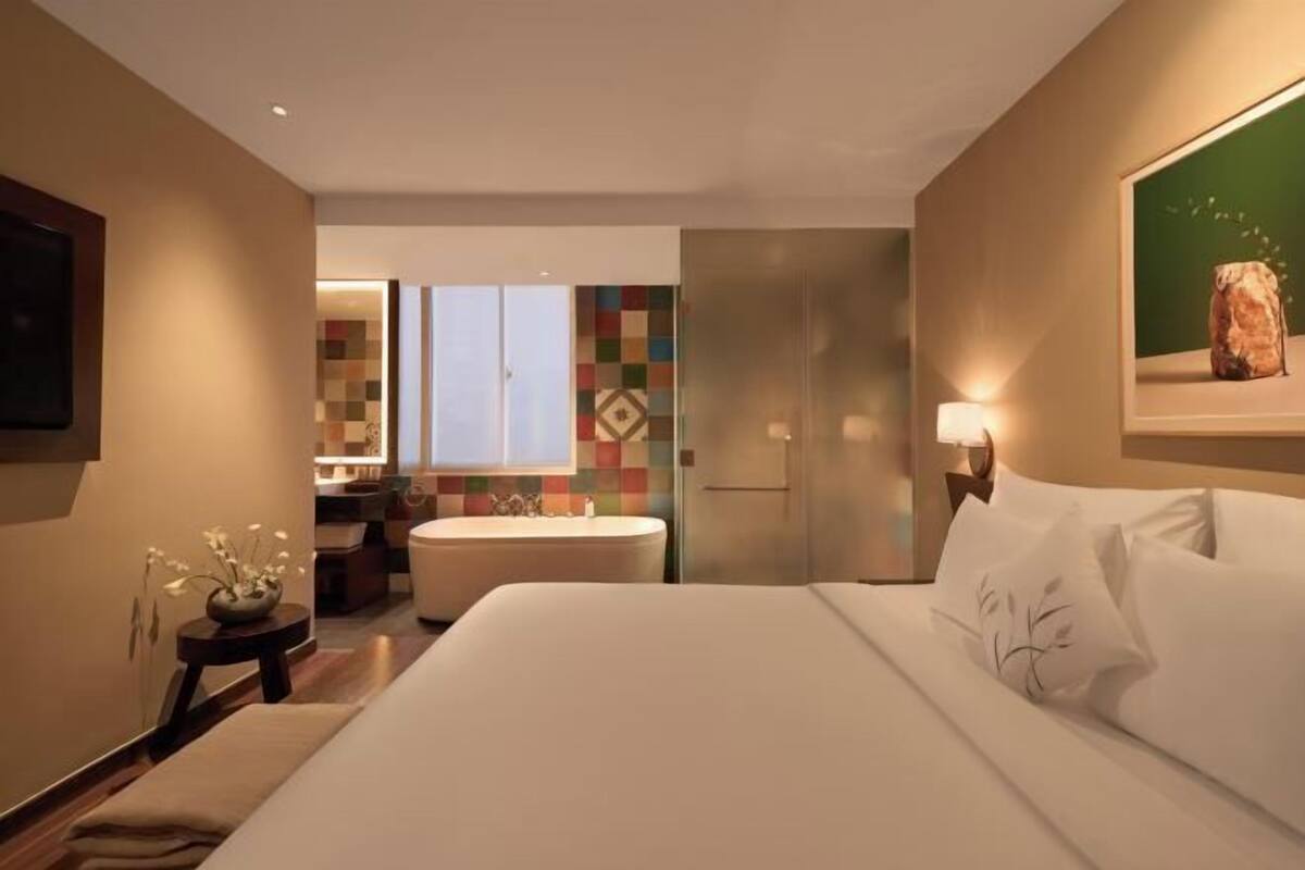 A hotel room with a bed, a nightstand, a lamp, a painting, and a bathroom with a bathtub and a window.