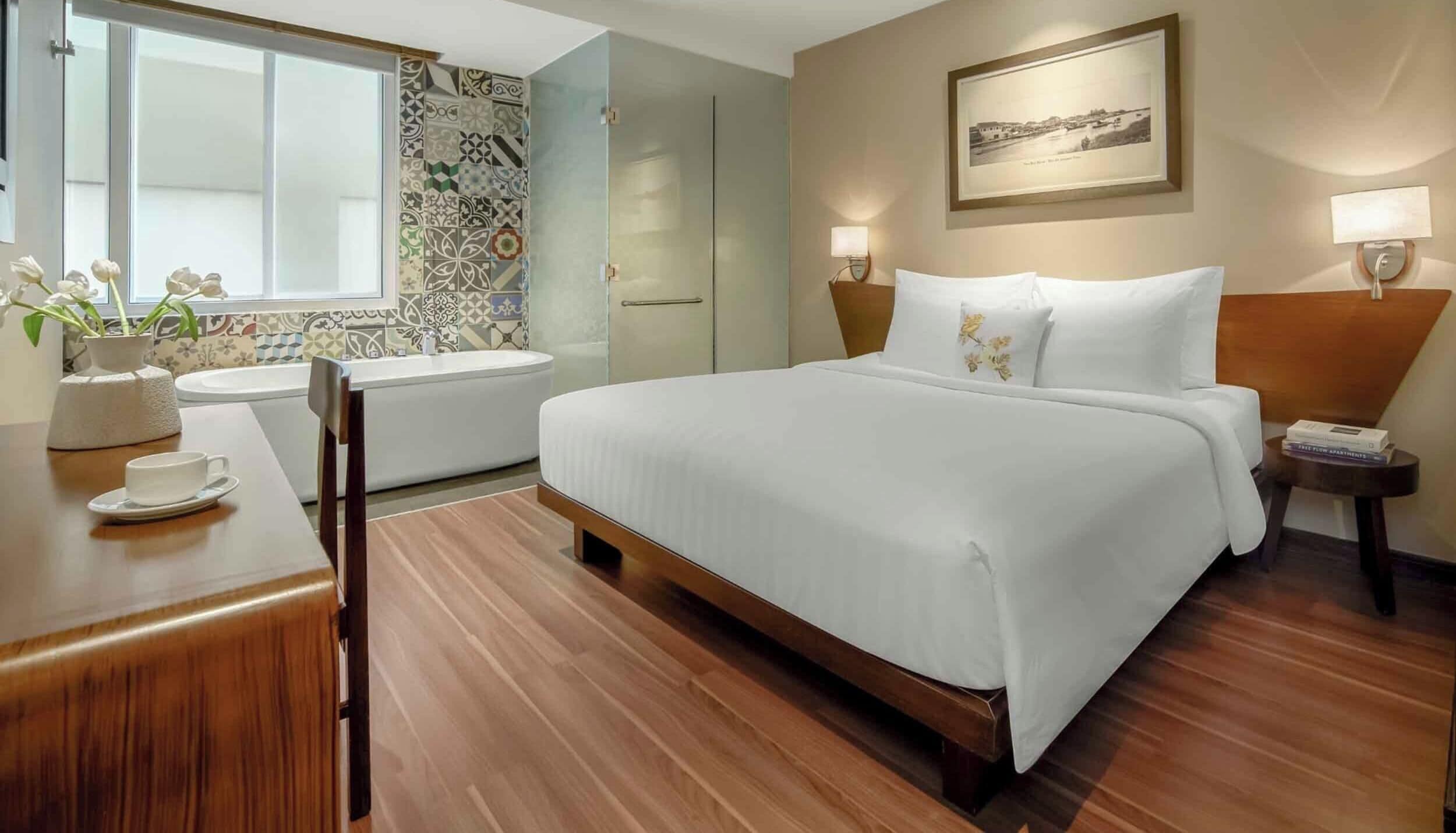 A hotel room with a bed, a desk with a cup and saucer, a window with flowers, a bathtub, and a wall with a picture.