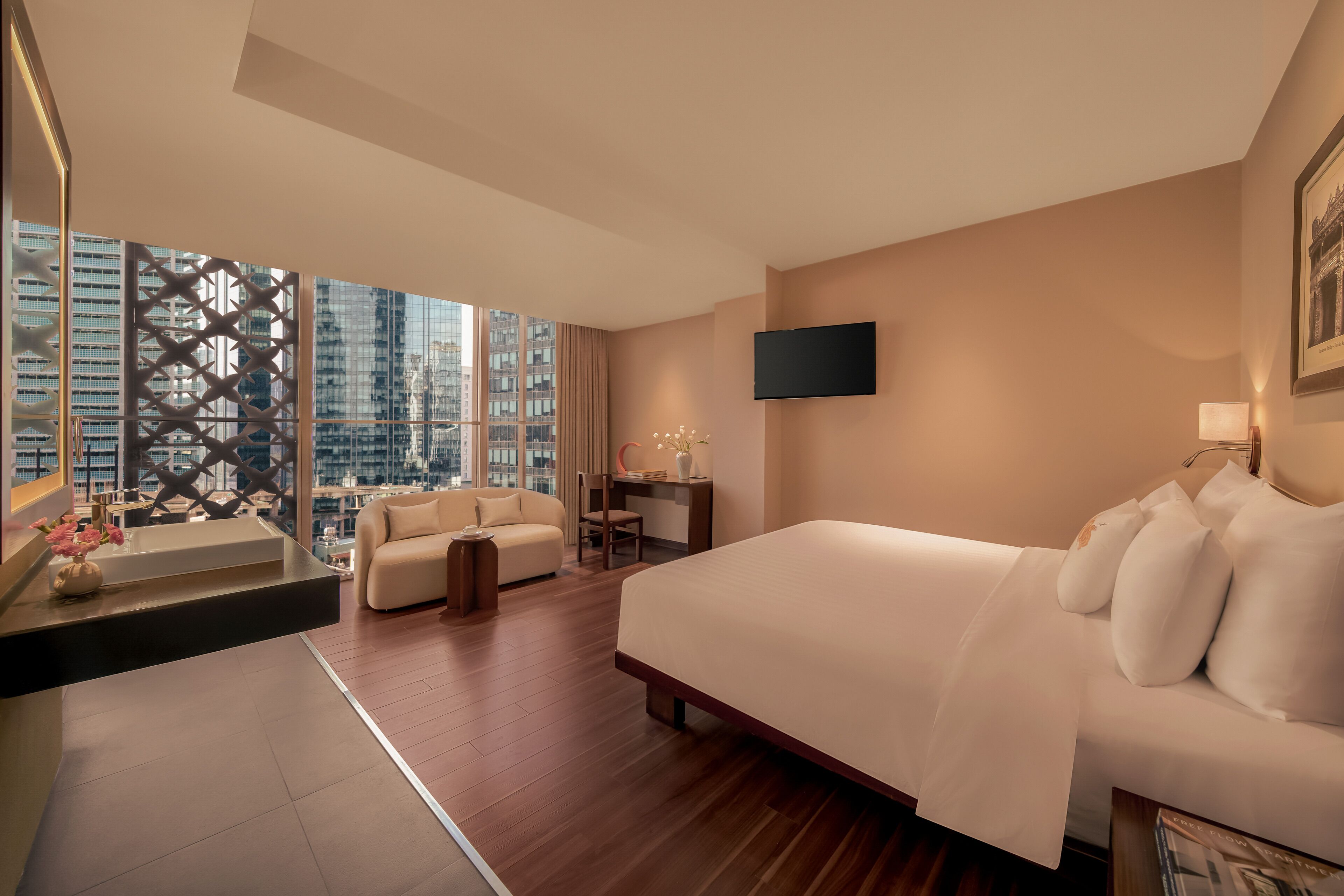 A modern hotel room with a large bed, a desk, a sofa, and a view of the cityscape through floor-to-ceiling windows.