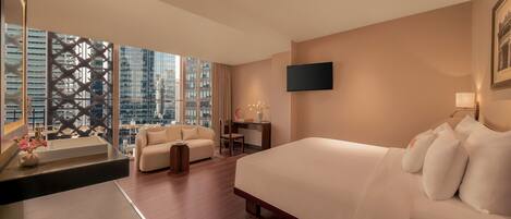 A modern hotel room with a large bed, a desk, a sofa, and a view of the cityscape through floor-to-ceiling windows.