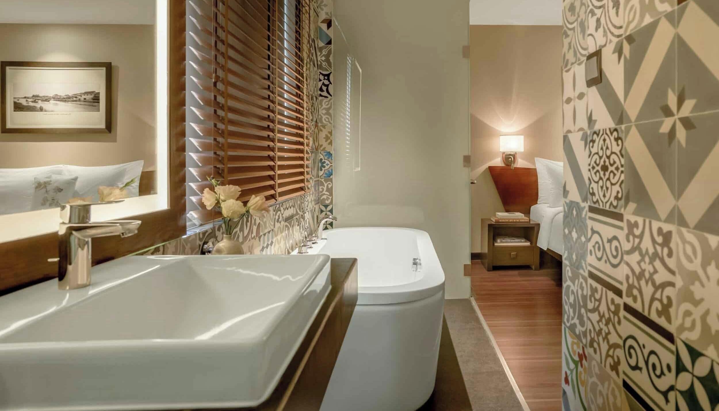 A bathroom with a large bathtub, a toilet, a mirror, and a small bedside table with a lamp.