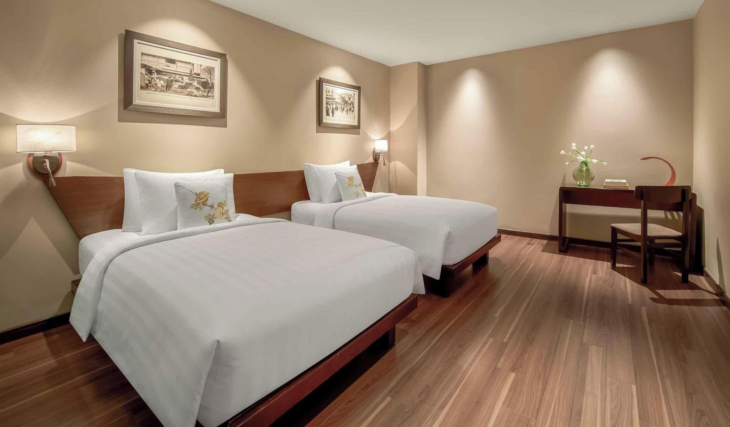 A hotel room with two beds, wooden flooring, wall-mounted lamps, and framed pictures.