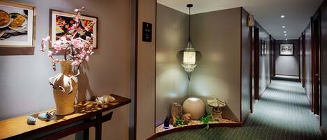 A hotel hallway with a wooden reception desk, a vase with pink flowers, a hanging light fixture, and a carpeted floor.