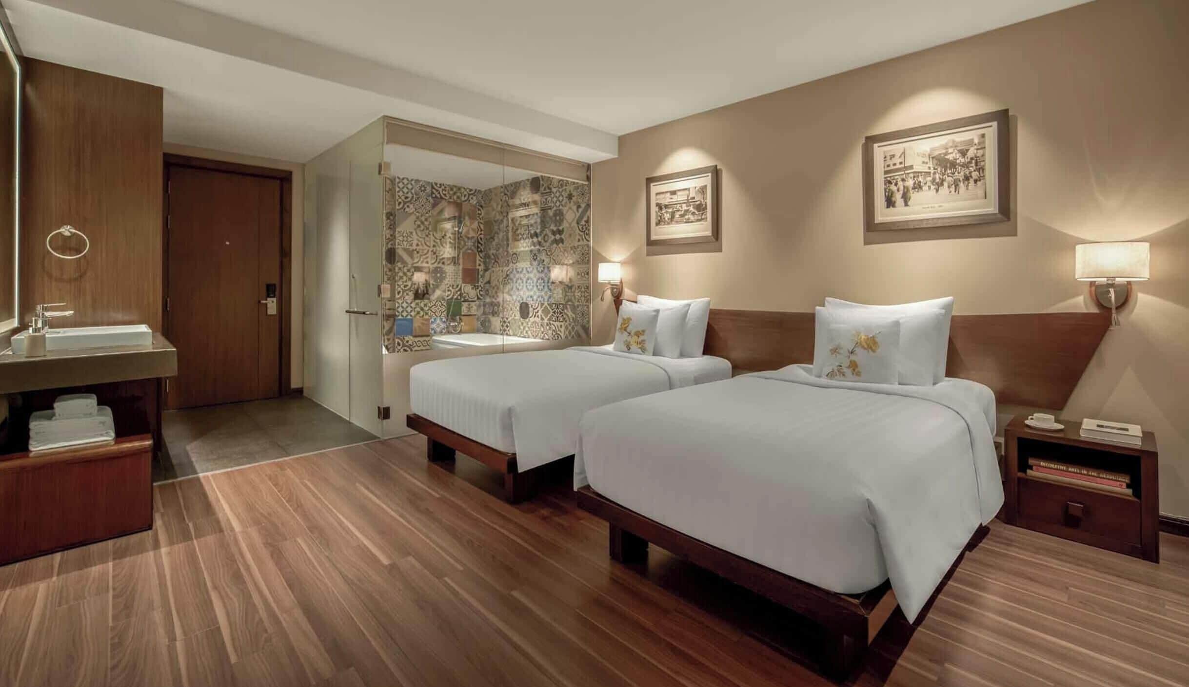 Ein Hotelzimmer mit zwei Betten, Holzboden, Badezimmer mit gemusterten Fliesen und gerahmten Bildern an den Wänden.