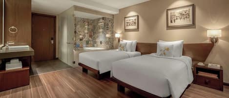 Ein Hotelzimmer mit zwei Betten, Holzboden, Badezimmer mit gemusterten Fliesen und gerahmten Bildern an den Wänden.
