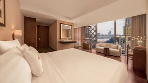 A hotel room with a large bed, a sitting area with a sofa and armchairs, a vanity with a mirror, and a view of the cityscape through large windows. - Silverland Sakyo Hotel & Spa (Ho Chi Minh City)