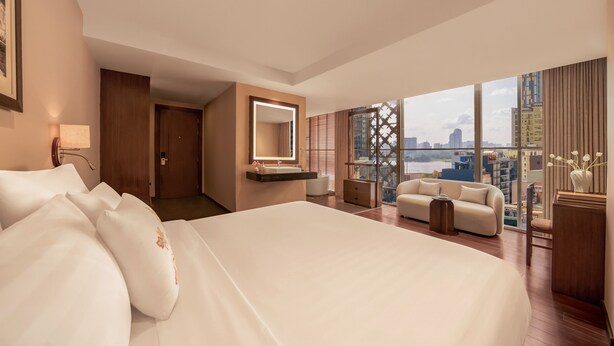 Habitación de hotel con una cama grande, una zona de estar con sofá y sillones, un tocador con espejo, y vistas a la ciudad a través de amplios ventanales.