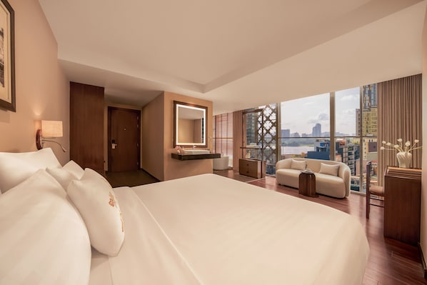 Habitación de hotel con una cama grande, una zona de estar con sofá y sillones, un tocador con espejo, y vistas a la ciudad a través de amplios ventanales.