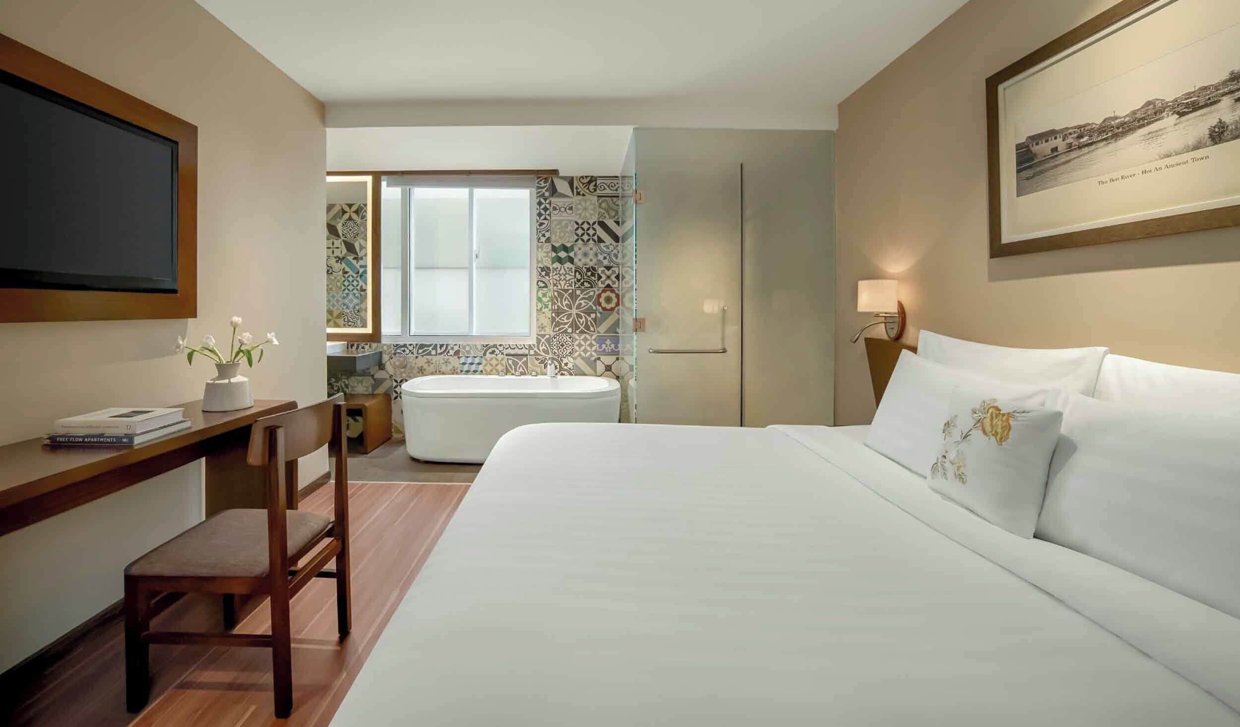 A hotel room with a bed, a desk, a chair, a TV, a bathroom with a bathtub, and a wall with a framed picture.