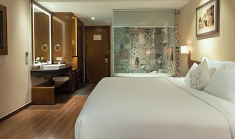 Habitación de hotel con cama, baño con ducha, tocador con espejo y un cuadro en la pared.