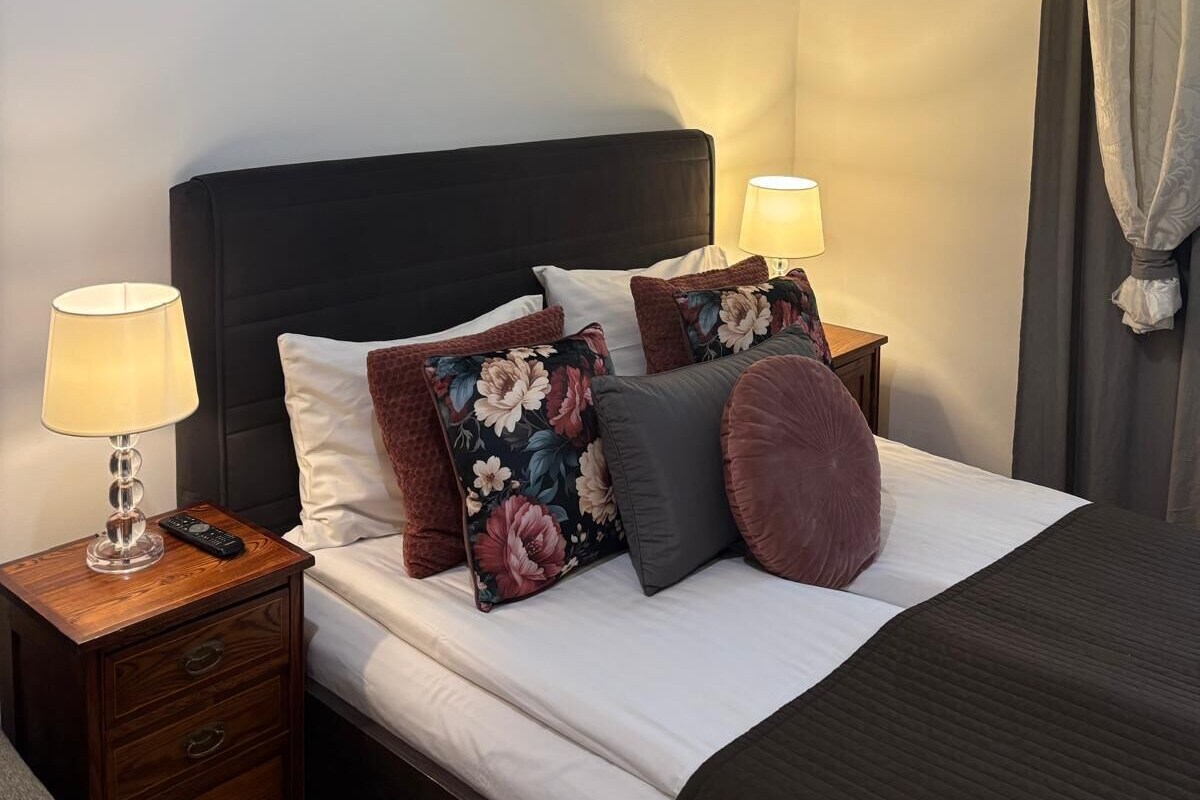 A neatly made bed with floral pillows, a bedside table with a lamp, and a wall-mounted picture frame.