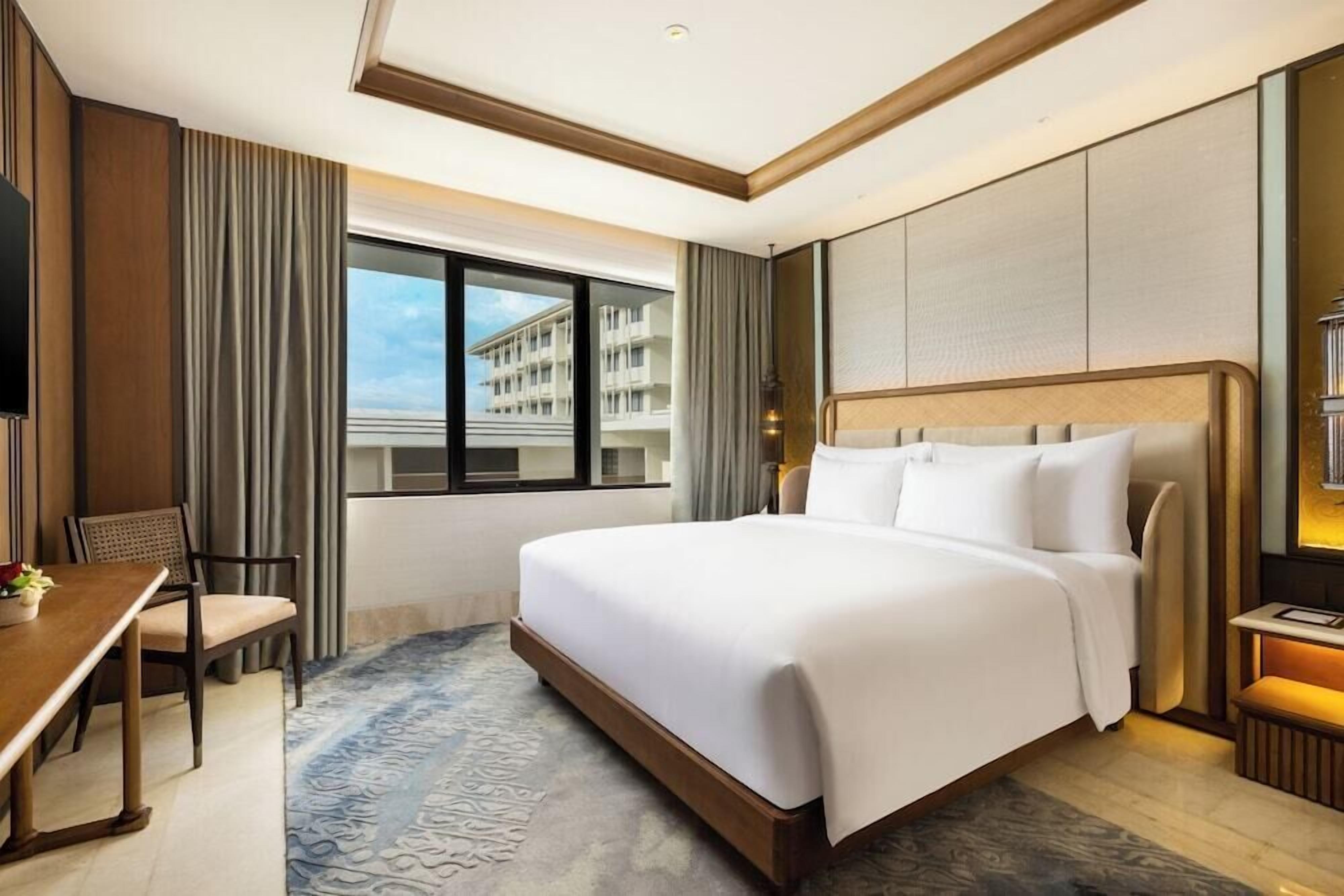 A hotel room with a large bed, a desk, and a chair. There is a window with a view of buildings.