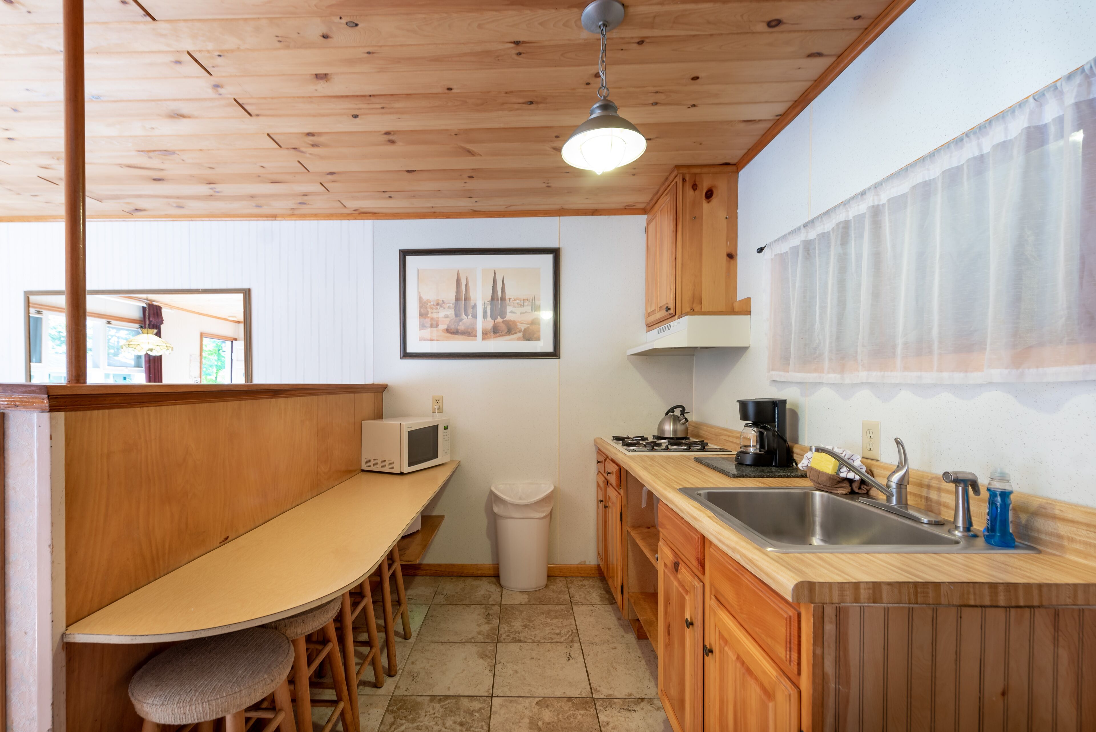 A compact kitchen with wooden cabinets, a microwave, a sink, and a small dining area with stools.