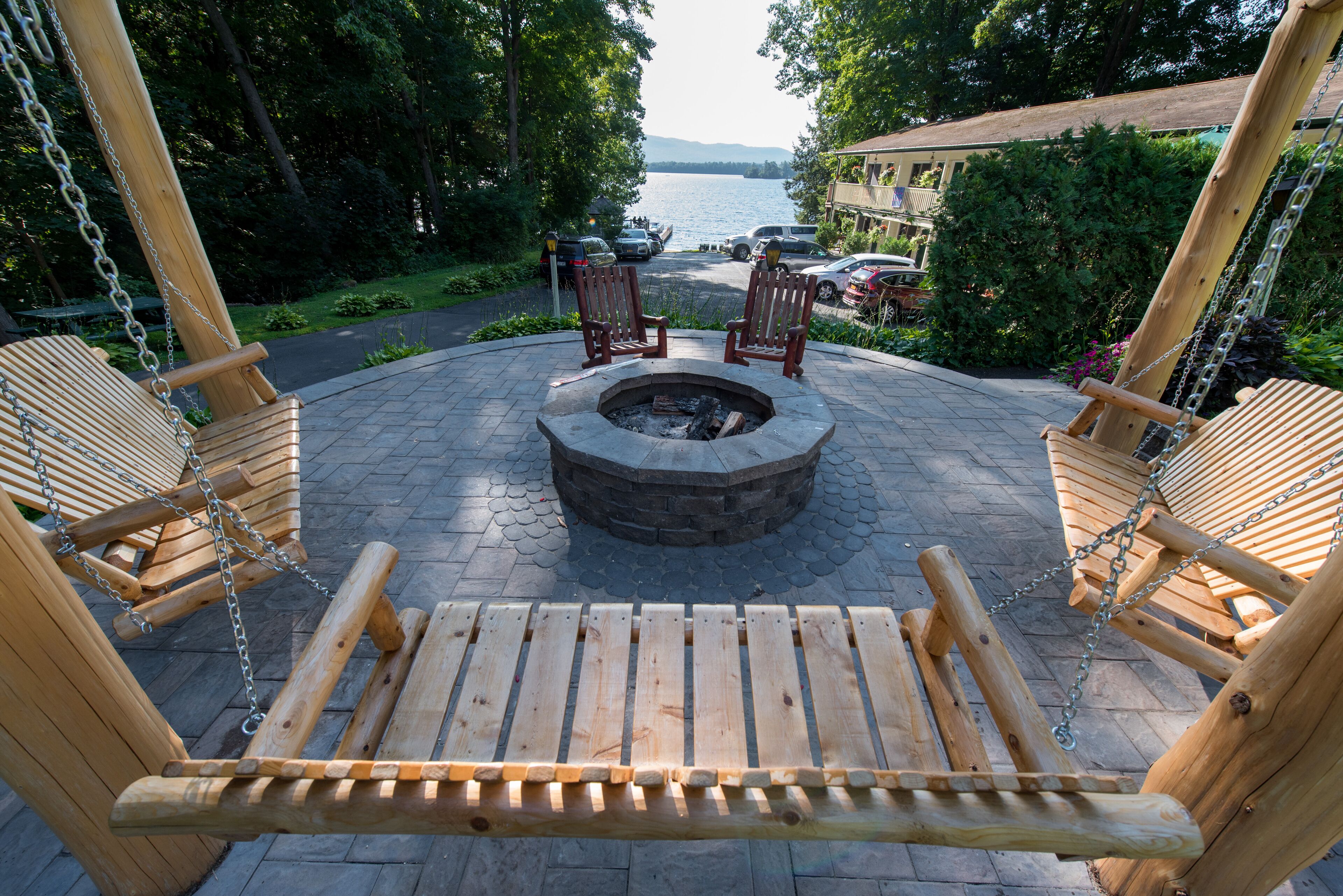A wooden swing set with a fire pit in the center, surrounded by a paved patio area with a view of a lake and parked cars in the background.
