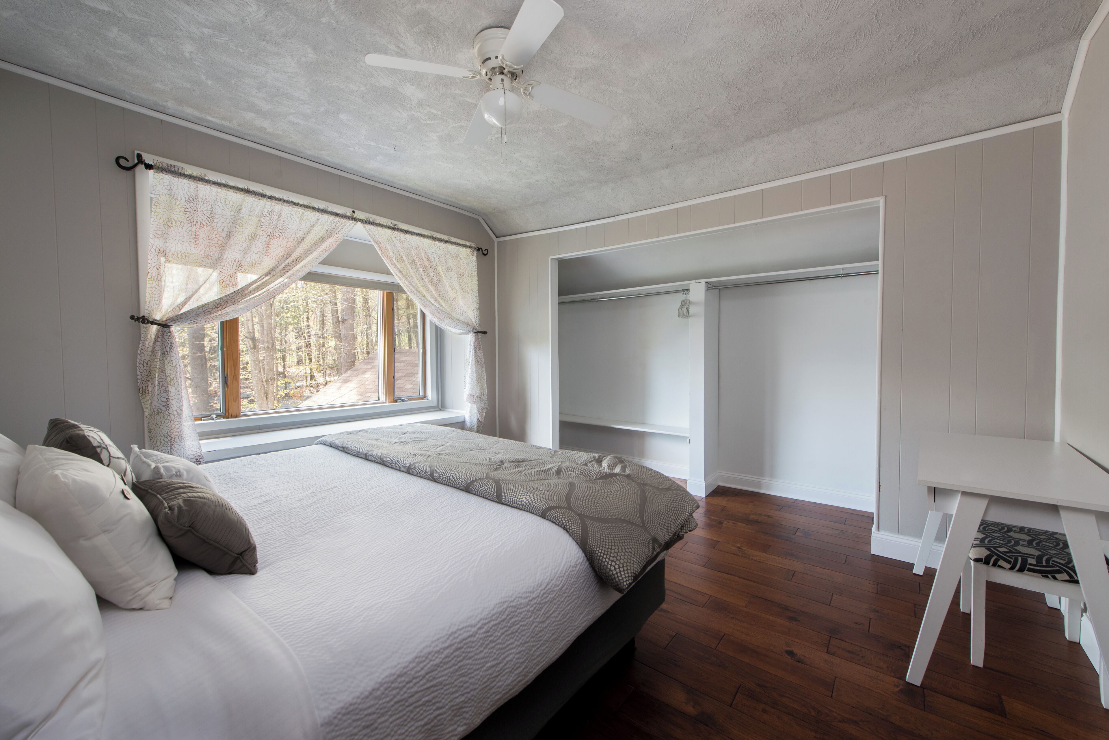 A bedroom with a bed, a ceiling fan, a window with curtains, a walk-in closet, and a wooden floor.