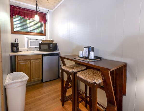 A compact kitchen with a microwave, coffee maker, and a small refrigerator. There are two wooden bar stools and a trash bin.
