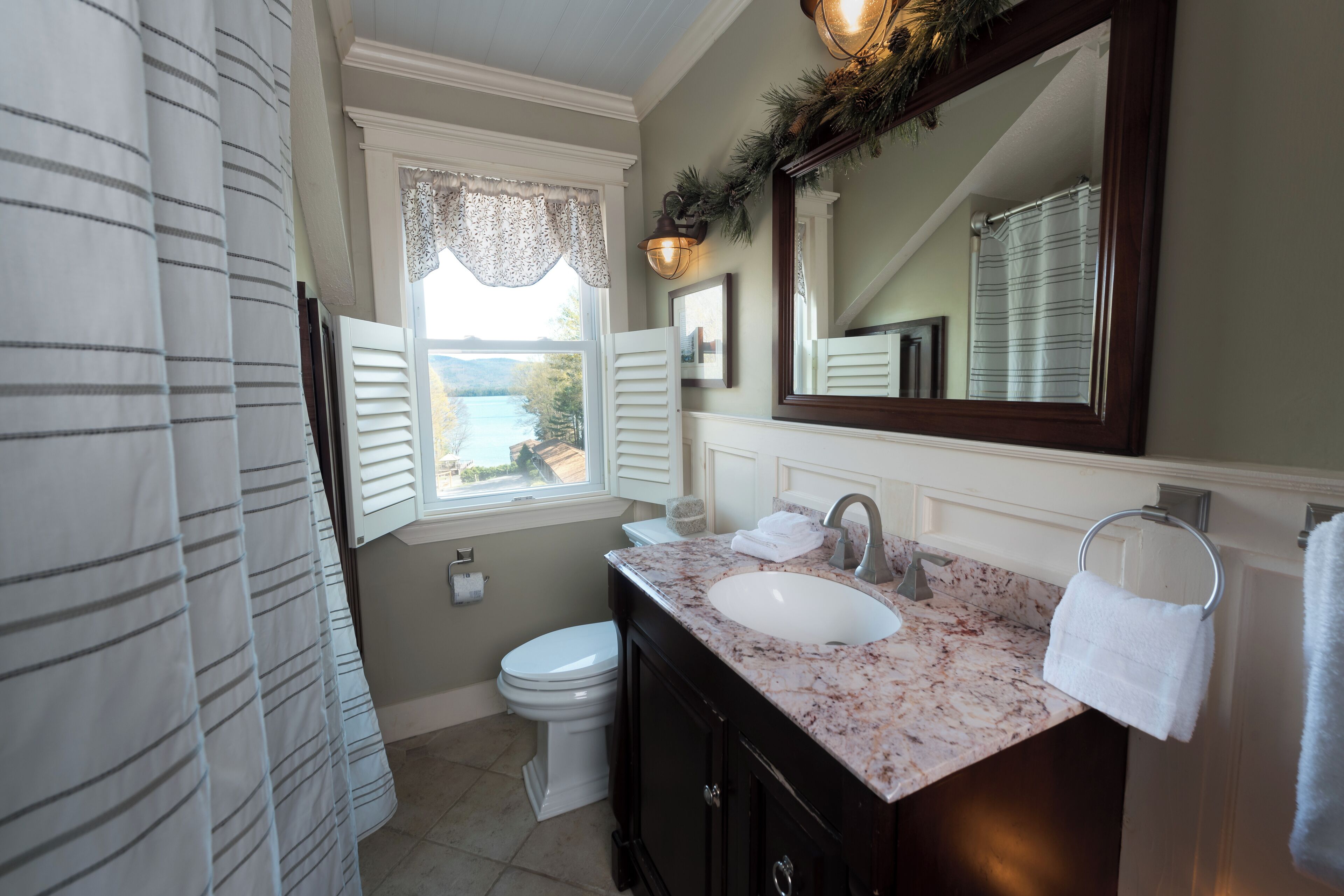A bathroom with a white toilet, a sink with a marble countertop, a large mirror, and a window with shutters.