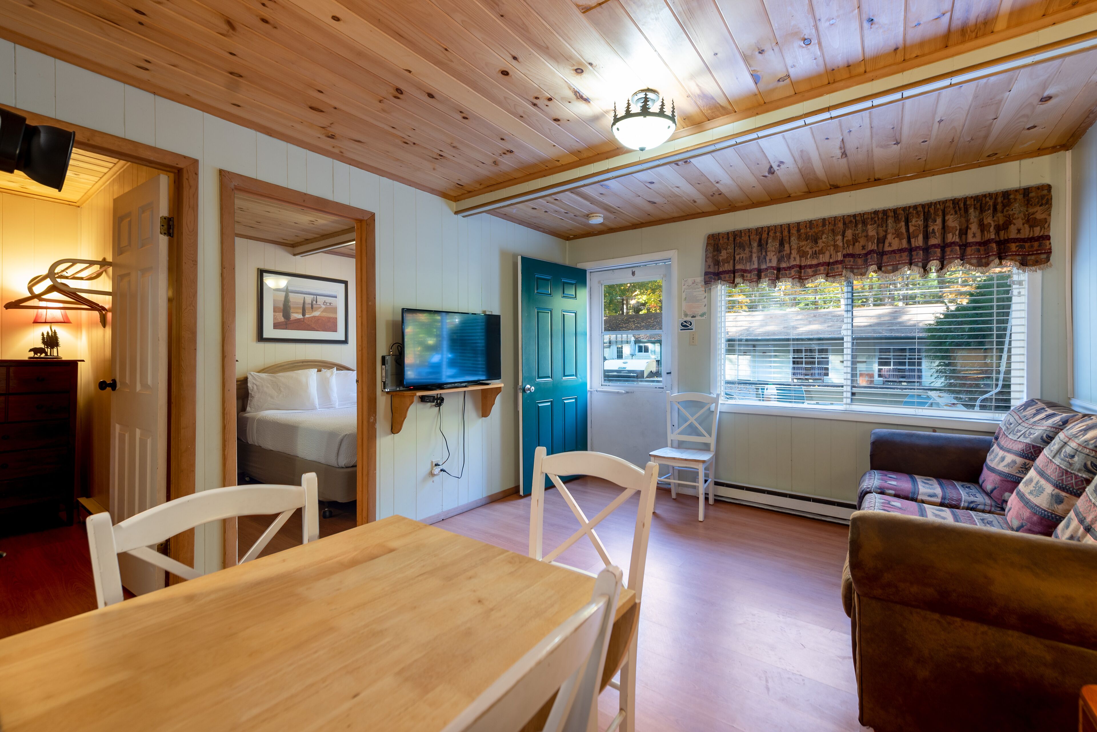 A cozy room with a wooden ceiling, a dining table with chairs, a sofa, a television, and a window with blinds.