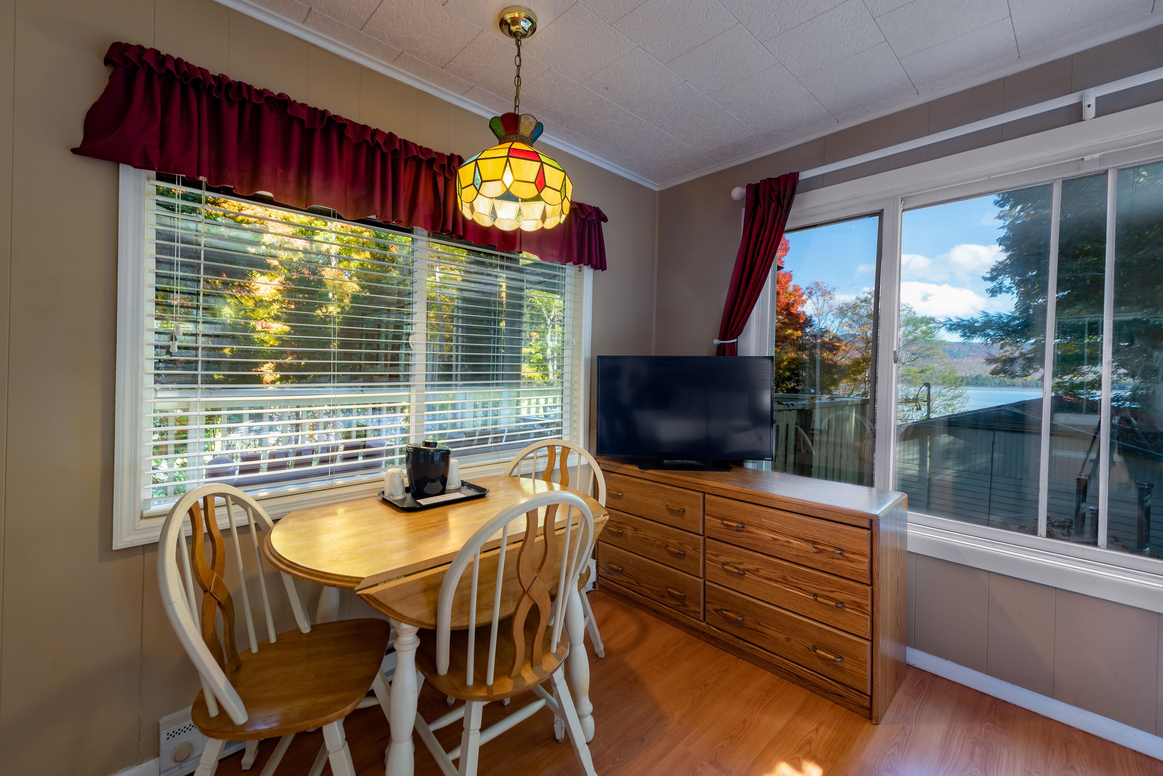A dining area with a wooden table and chairs, a television on a cabinet, and large windows with blinds.