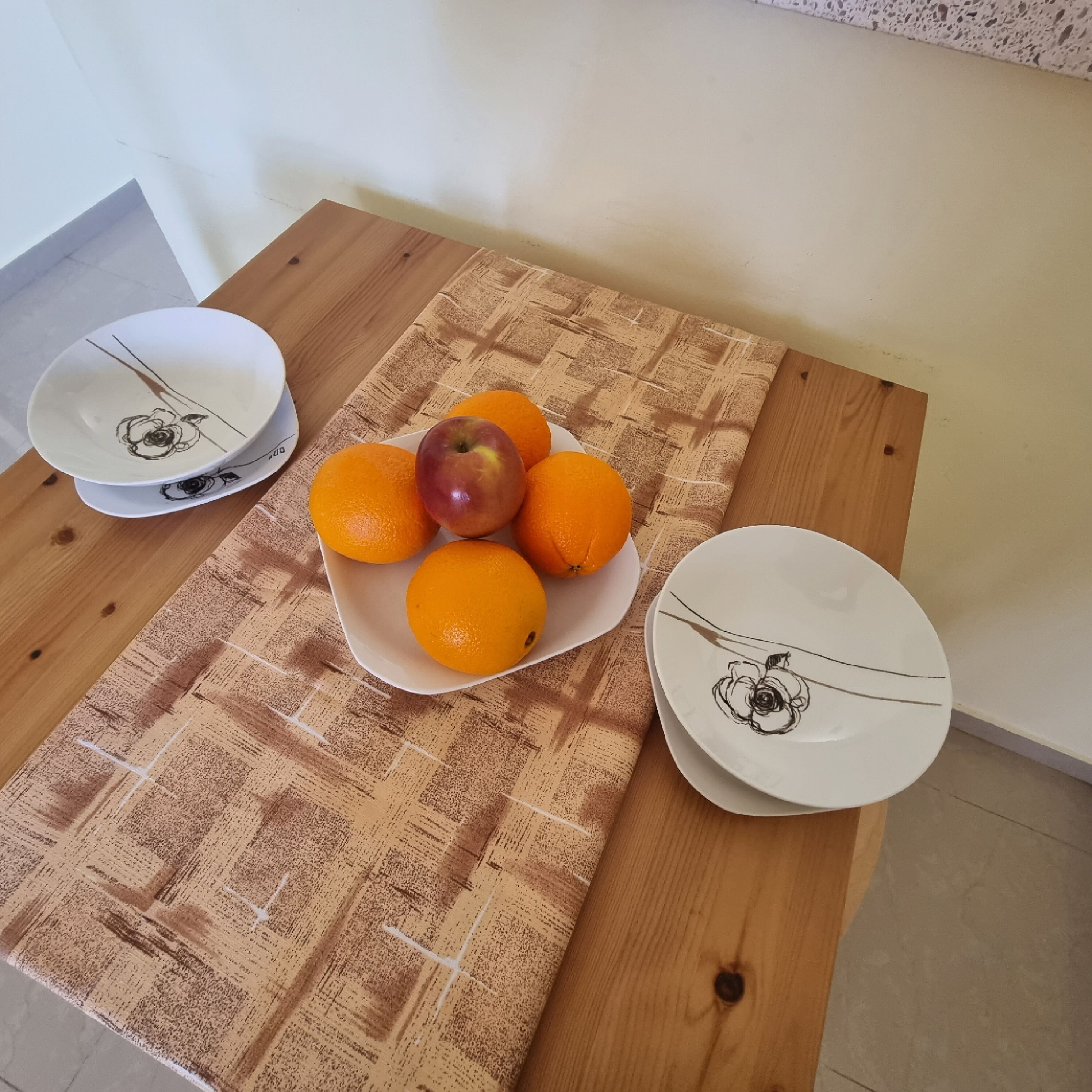 Uma mesa de madeira com toalha estampada, uma tigela de laranjas e uma maçã, e duas travessas com desenho de rosa.