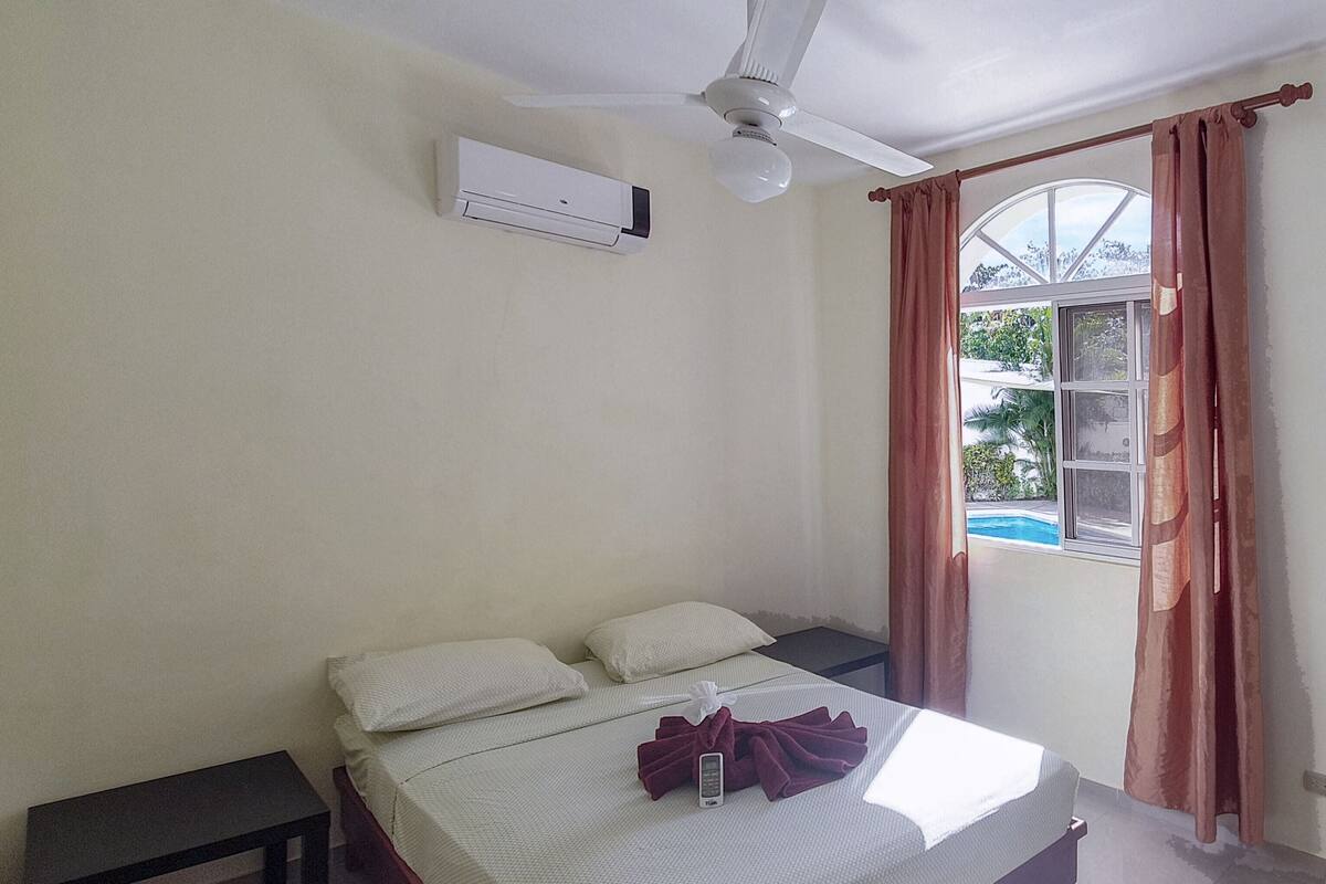 A single bed with a white comforter, a bedside table, an air conditioning unit, a ceiling fan, and a window with curtains.