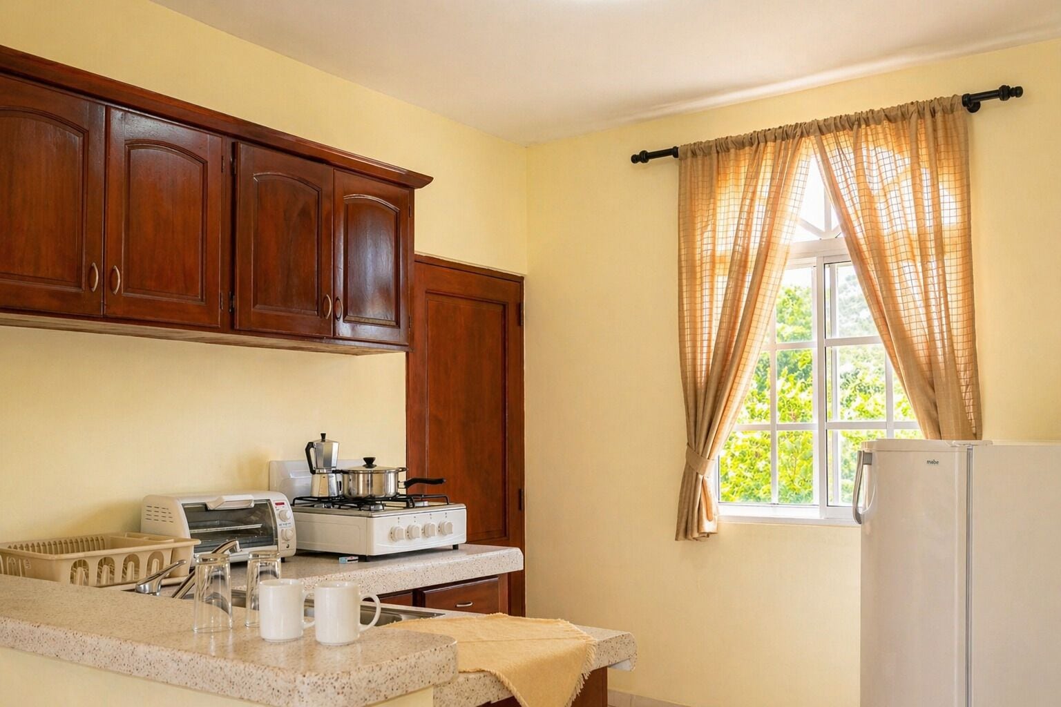 Cucina con mobili in legno, piano cottura a gas, frigorifero bianco e finestra con tende beige.