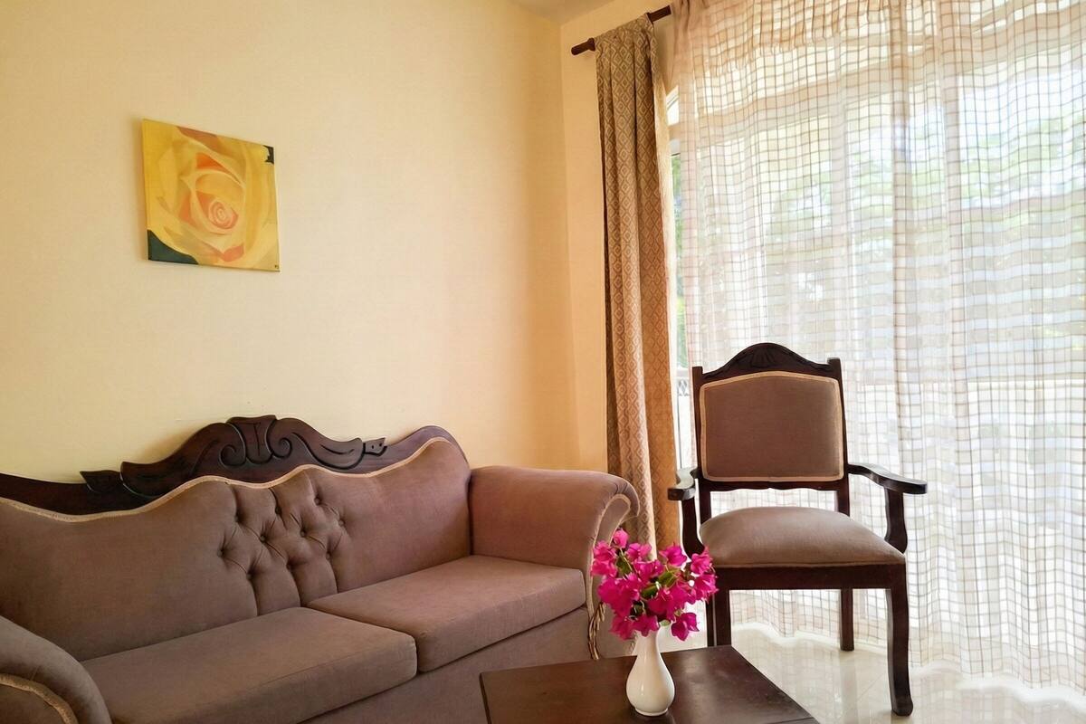 A living room with a brown sofa, a wooden chair, a vase with flowers, and a painting on the wall.