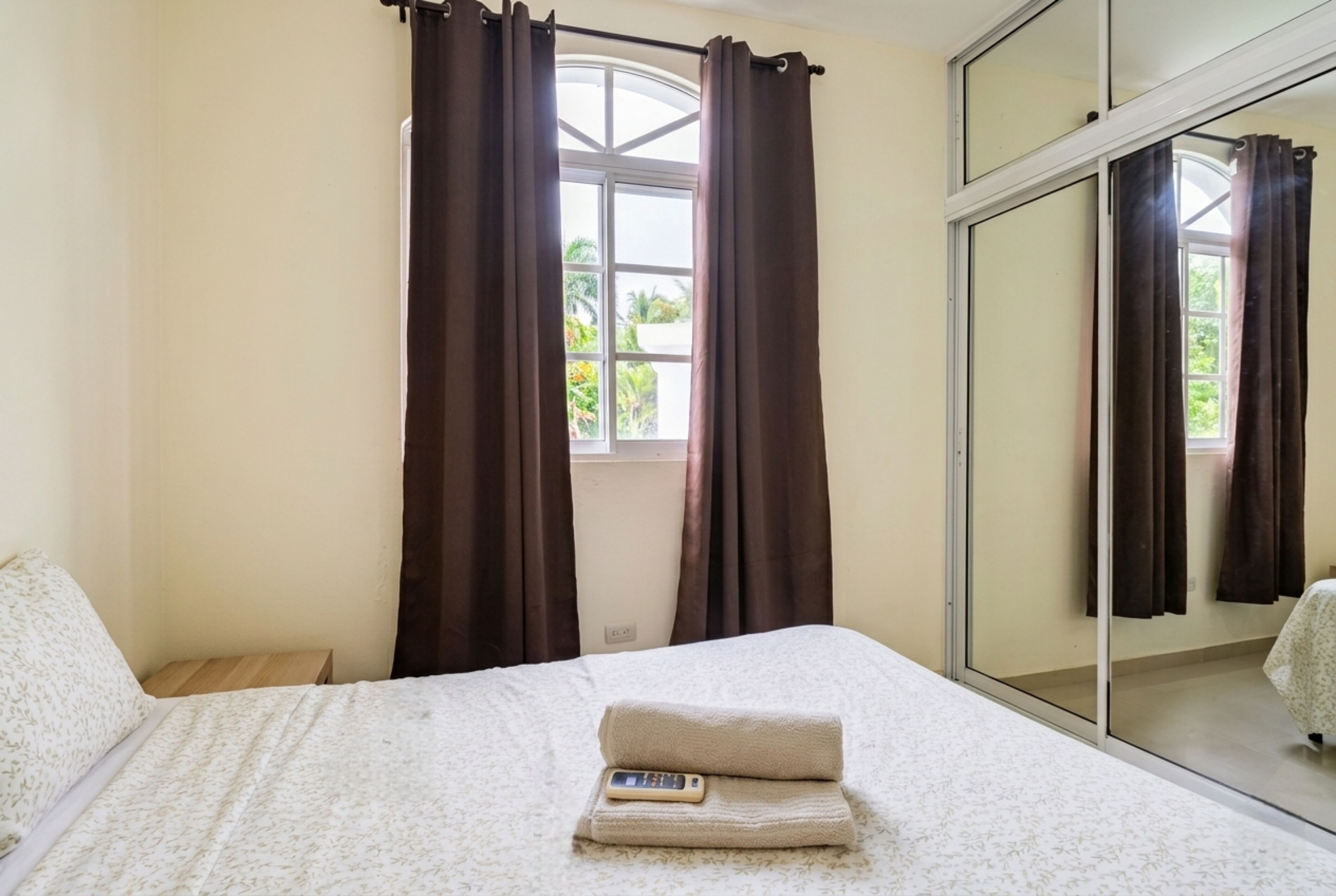 A bedroom with a bed, a window with curtains, a mirror, and a view of greenery outside.