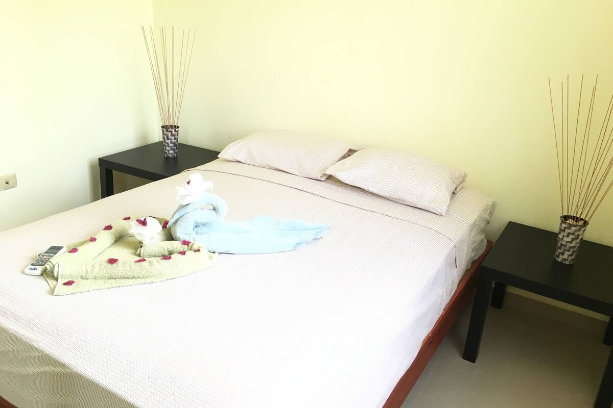 A neatly made bed with a towel and a decorative item, two bedside tables with vases, and a plain wall.
