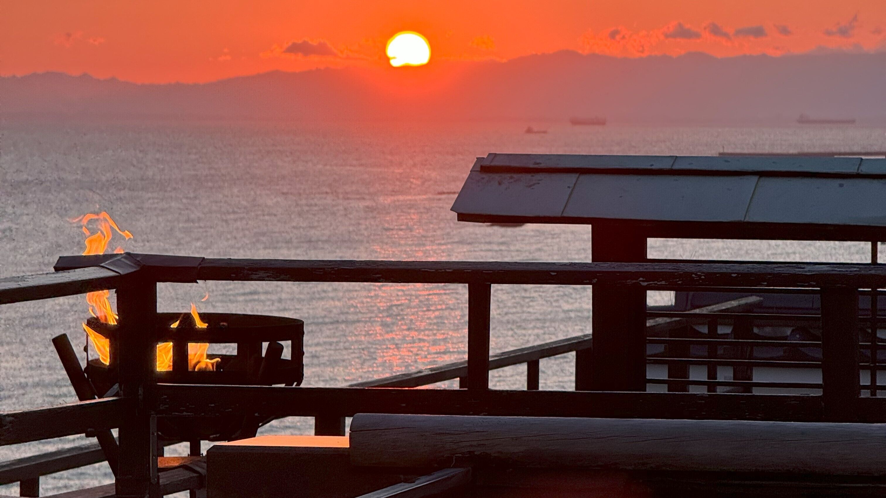 A wooden pier with a grill and a table, overlooking a body of water with boats and a sunset in the background.