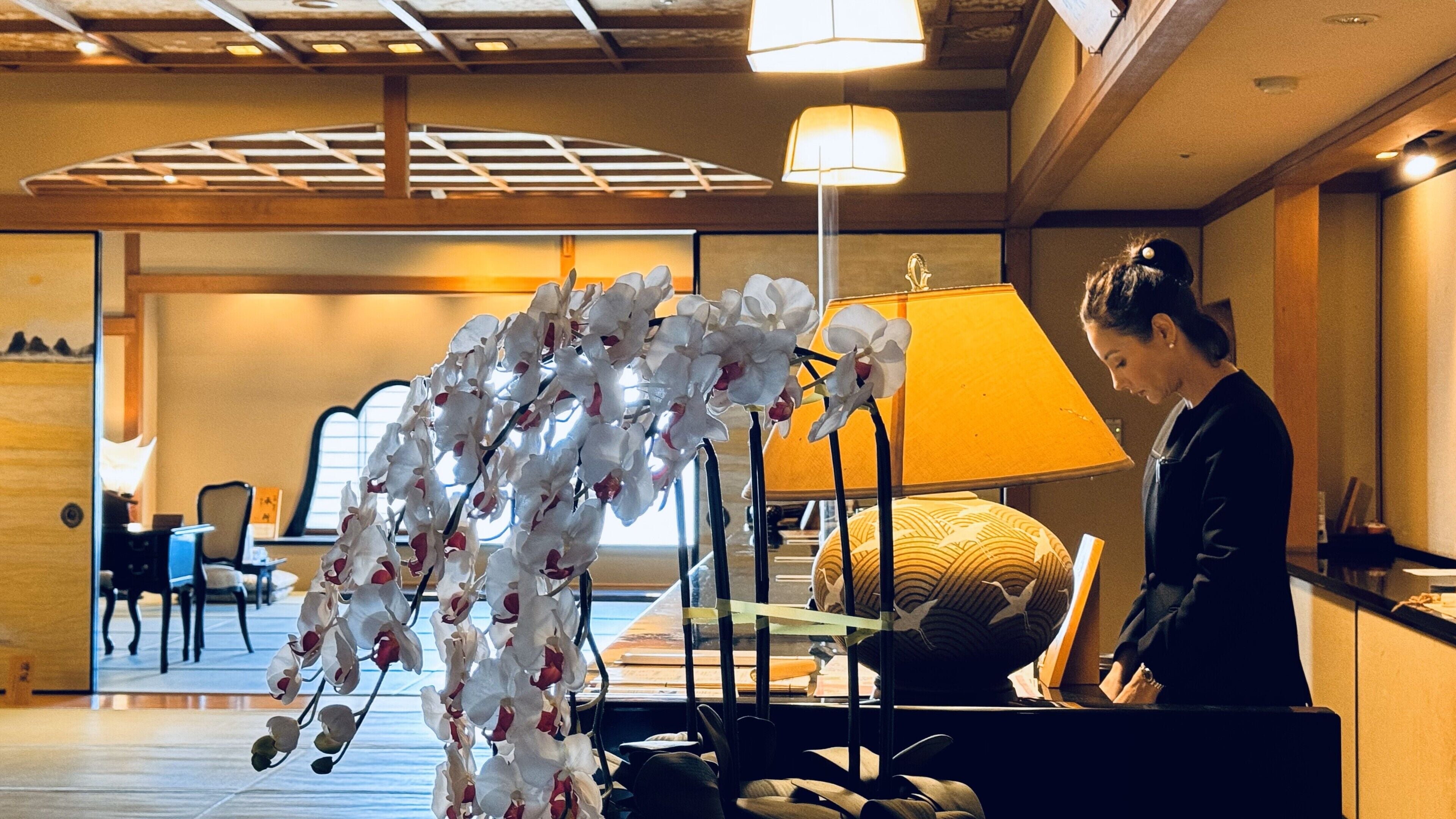 A hotel room with a large orchid arrangement, a bed with a patterned pillow, a desk with a lamp, and a view of the room's interior.
