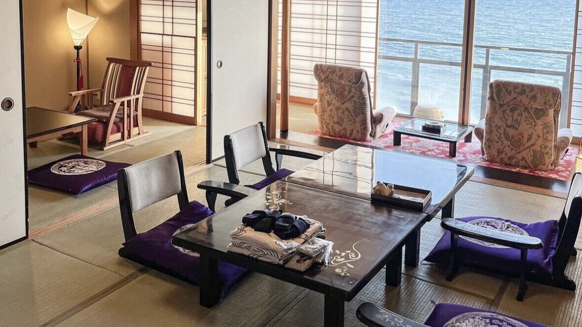A traditional Japanese-style room with tatami flooring, a low table with cushions, and a view of the sea through large windows.