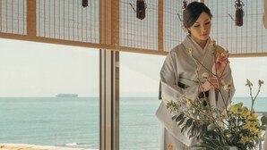 Een kamer met een groot raam dat uitkijkt op de zee, een traditioneel Japans interieur en een bloeiende plant in een pot.