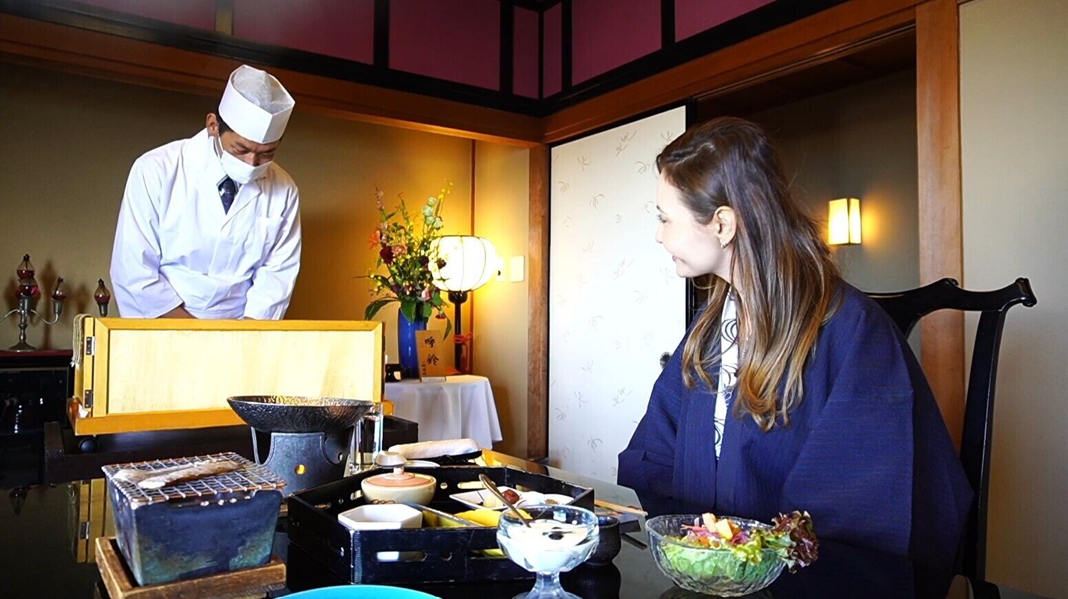 Uma sala de jantar com mesa posta para dois, um chef de uniforme e diversas refeições e utensílios.