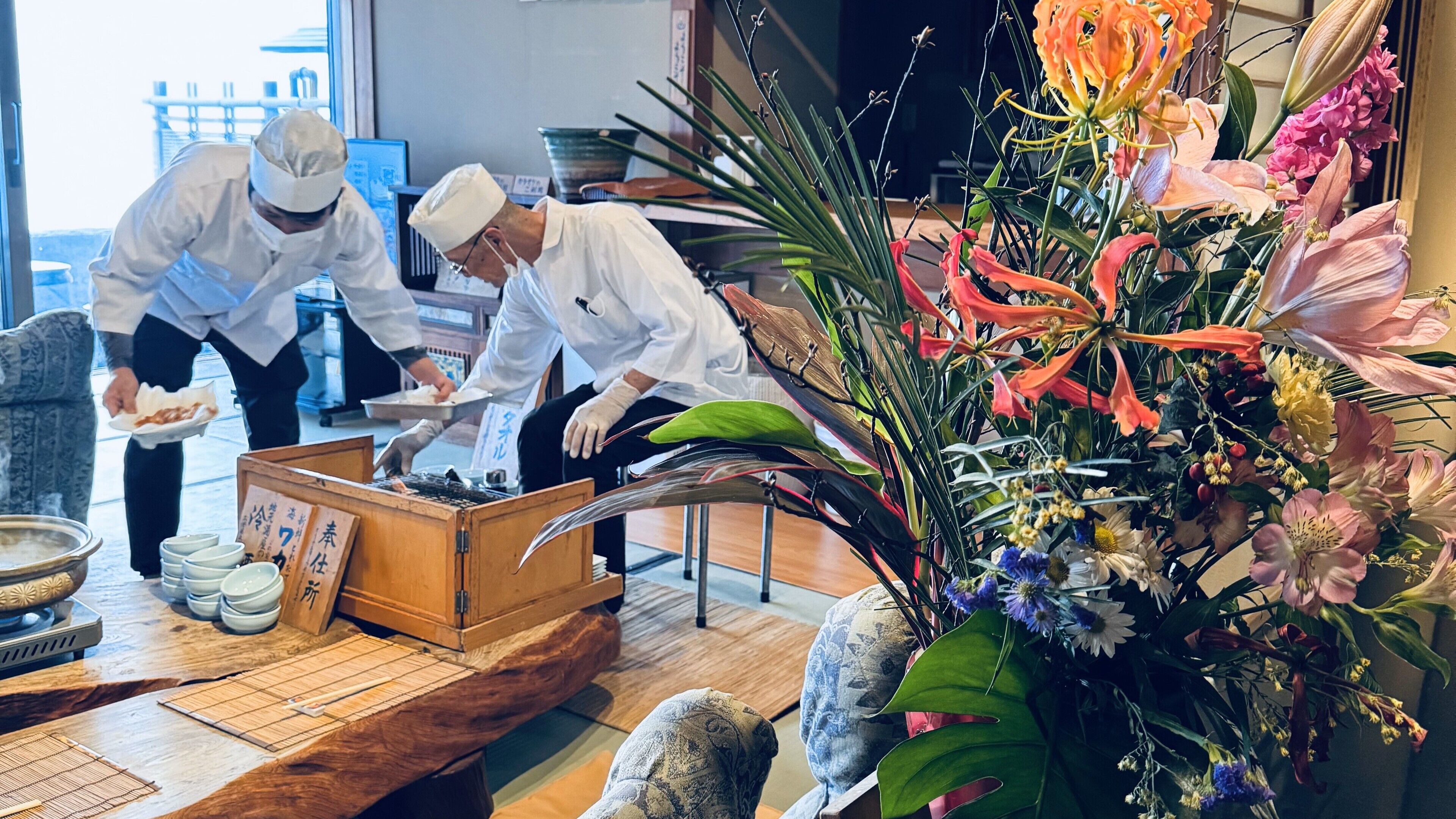 Interior de un restaurante mostrando a cocineros preparando comida, una mesa de madera con platos, y un arreglo floral grande.