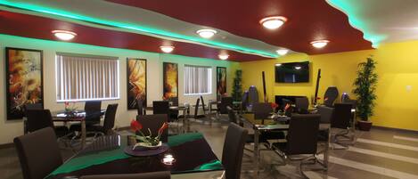 A conference room with multiple tables and chairs, a flat-screen TV mounted on the wall, and a large window with blinds.