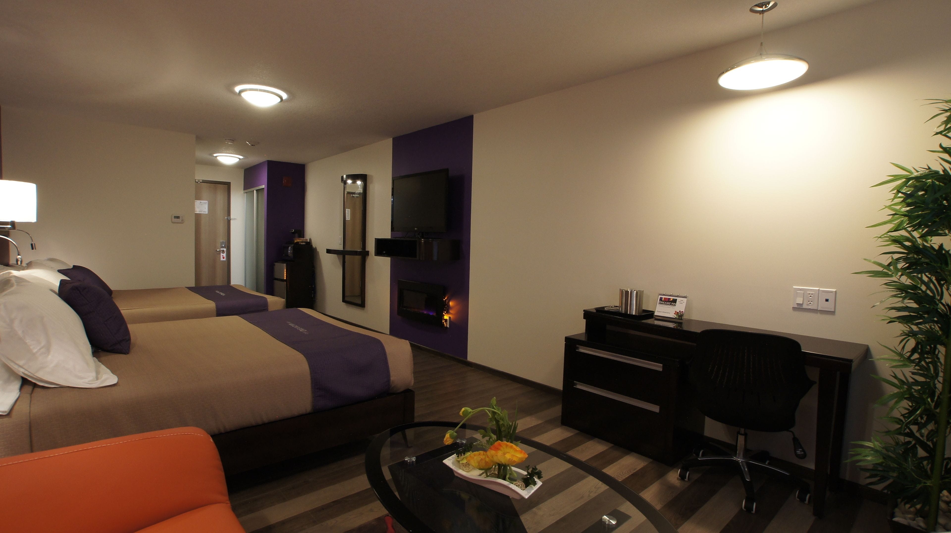 A hotel room with a bed, a desk, a chair, a small table with flowers, and a potted plant.