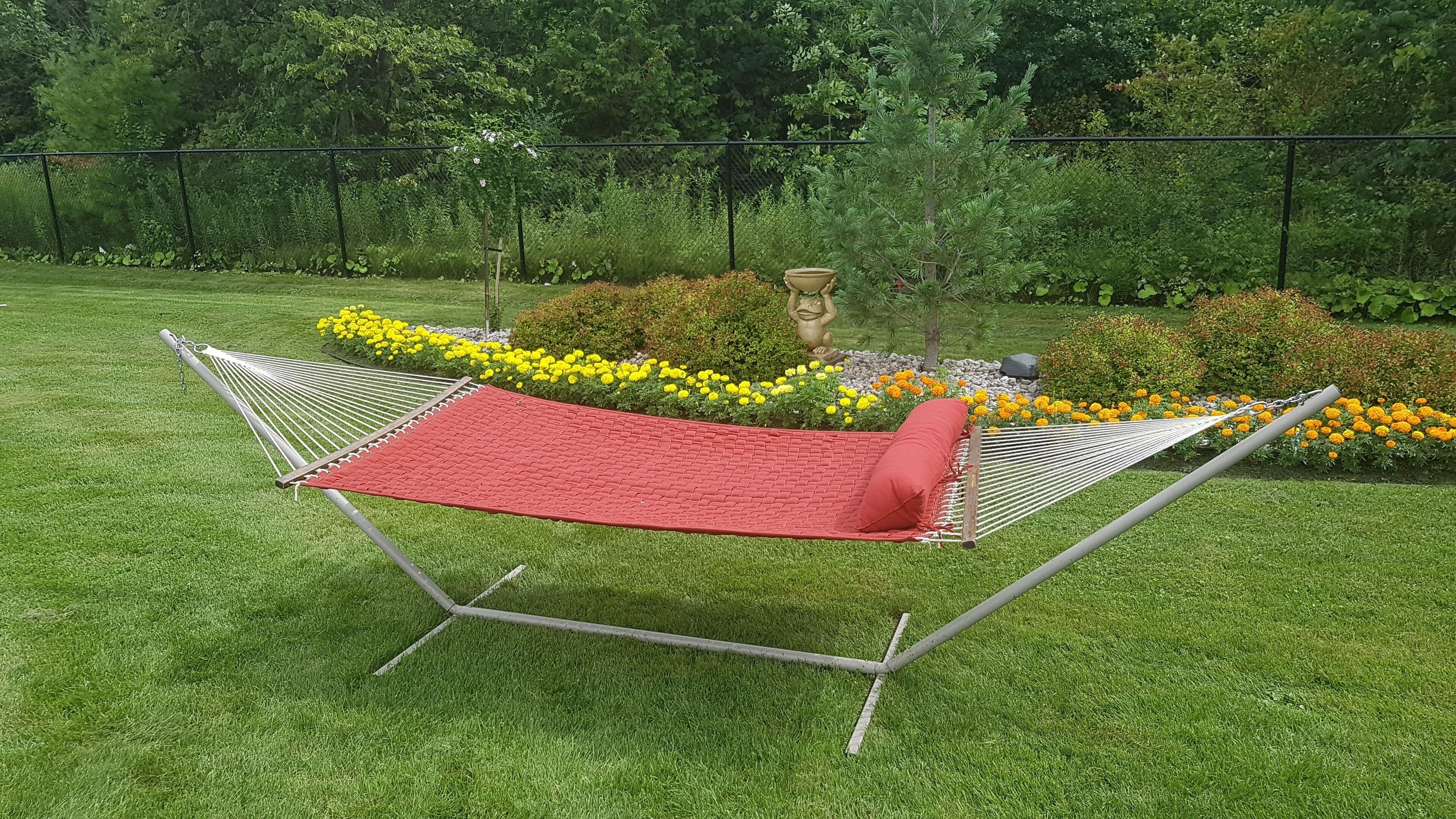 Een hangmat met een rode mat en een rood kussen, in een grasveld met gele bloemen en een standbeeld op de achtergrond.