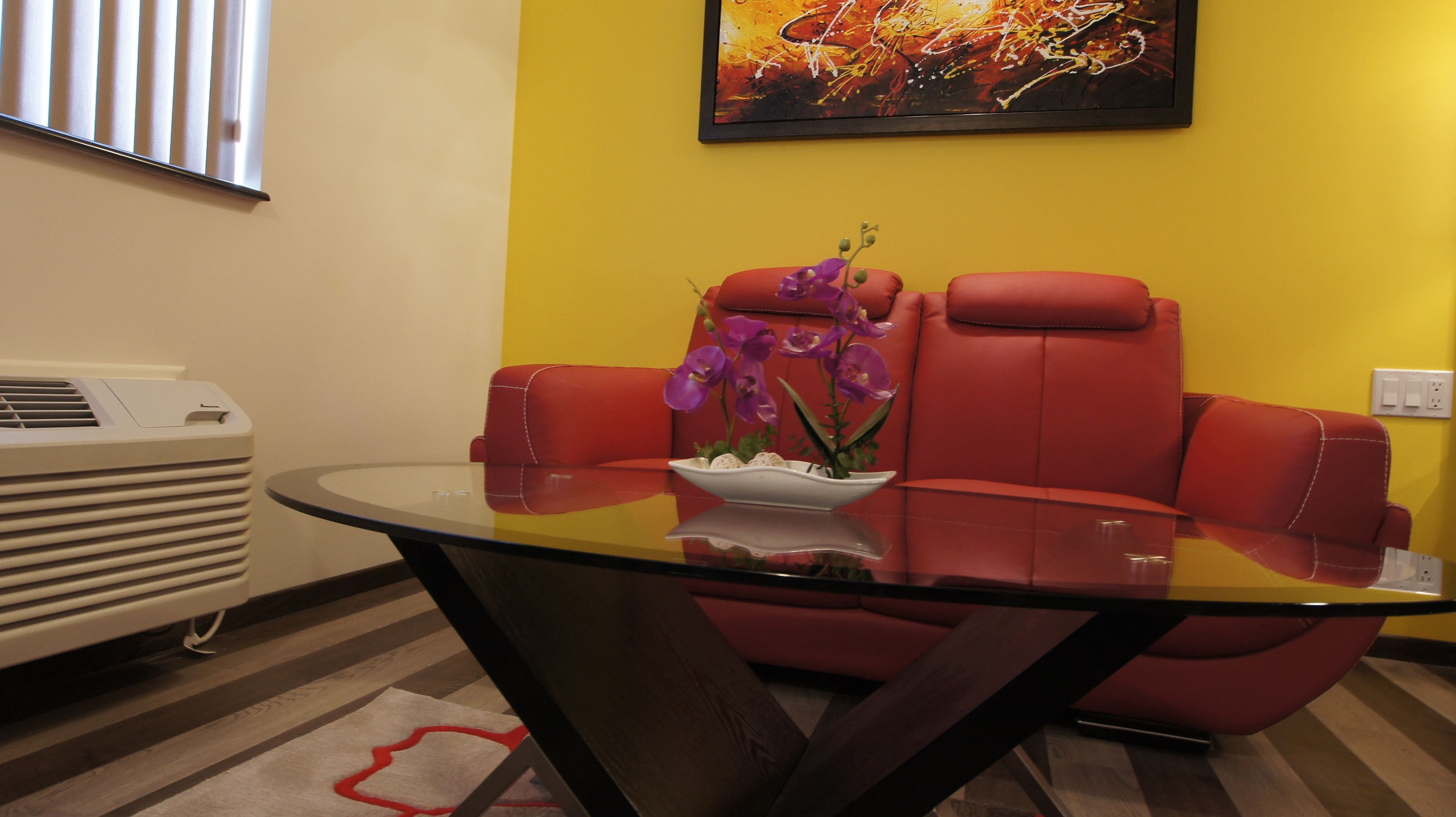 A modern room with a glass table, red leather chairs, a white air conditioner, and a vibrant yellow wall.