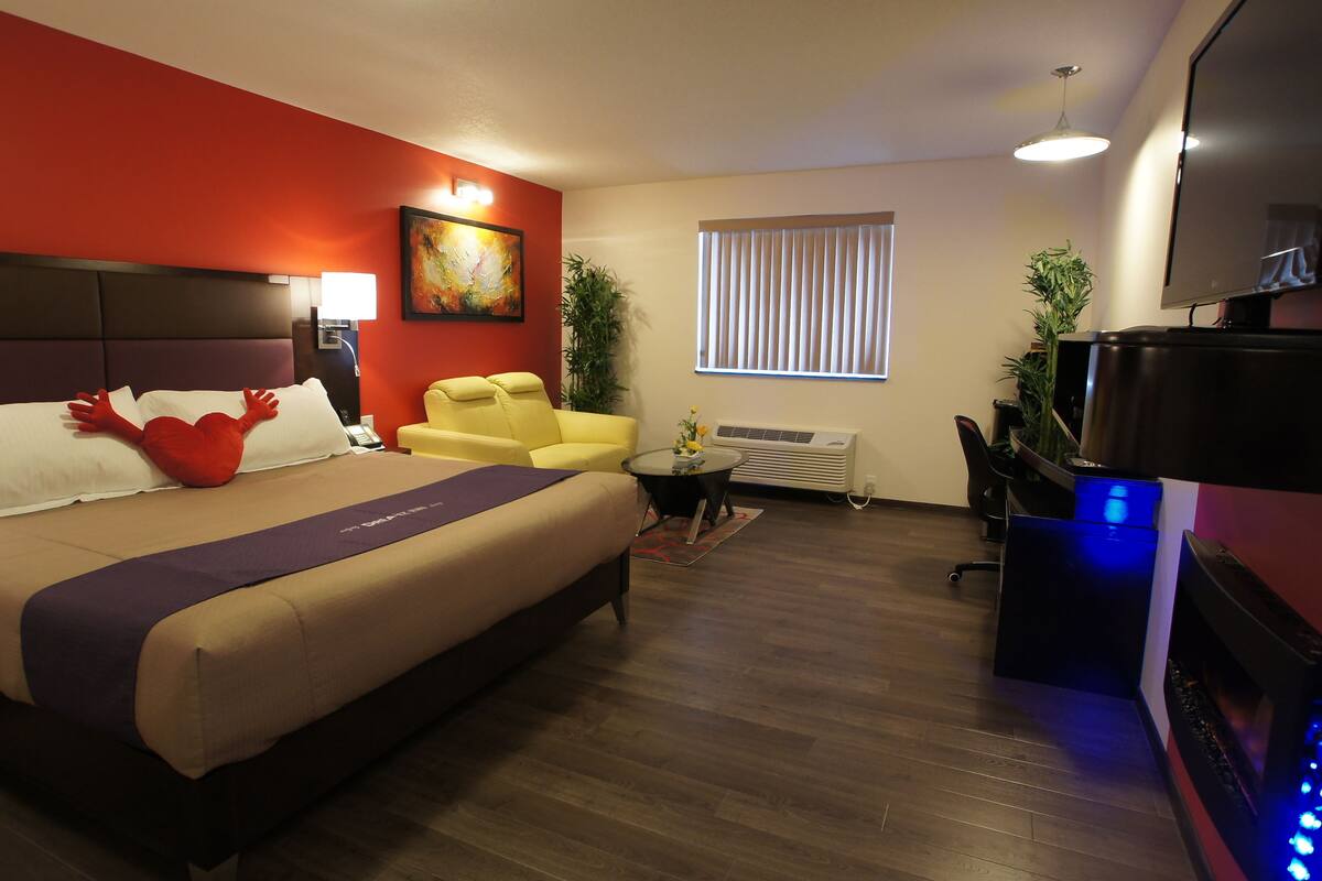 A hotel room with a bed, a yellow armchair, a desk with a computer, a TV, and a window with blinds.