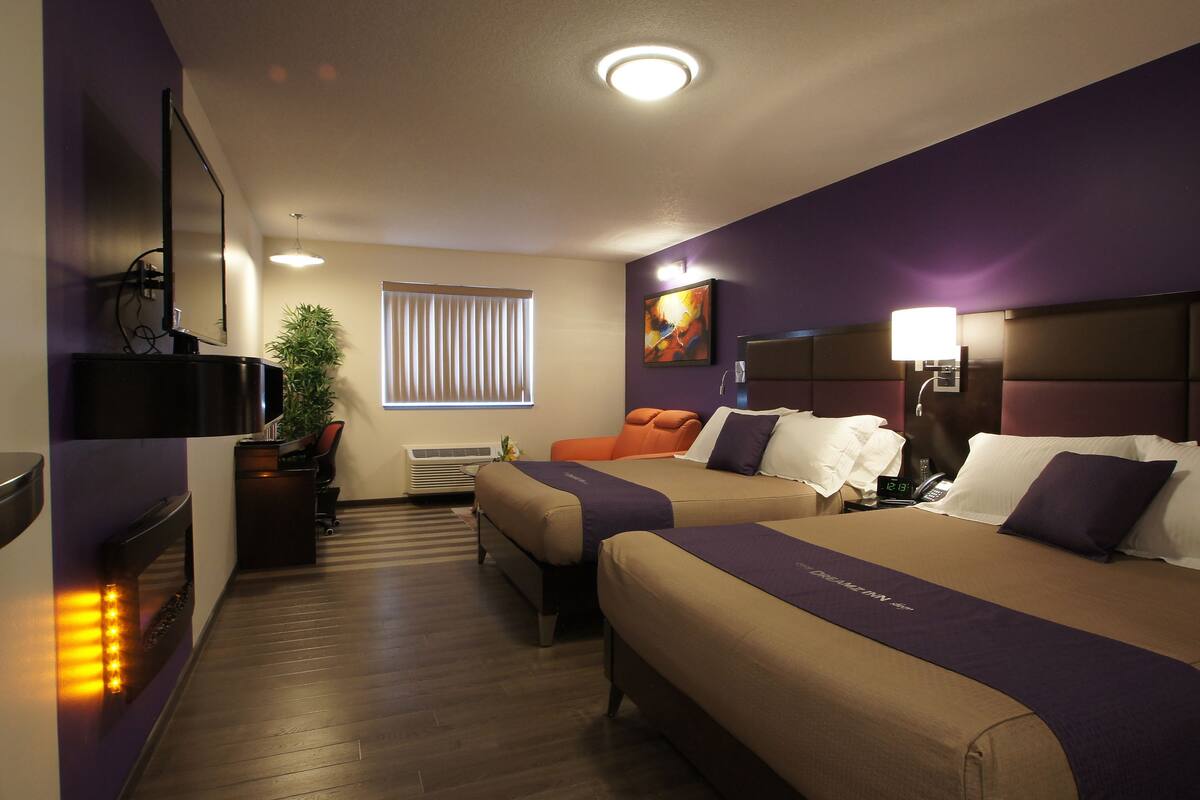 A hotel room with two beds, a desk, a chair, a plant, a window with blinds, and a painting on the wall.