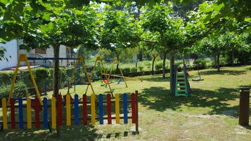 Un parque infantil con columpios, tobogán y estructura para escalar, rodeado de árboles y césped.