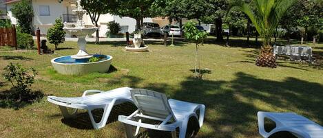 A grassy outdoor area with a fountain, white plastic chairs, and a small tree.