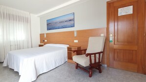 Habitación de hotel con una cama, una mesita de noche de madera, una silla, una puerta de madera y un cuadro en la pared.