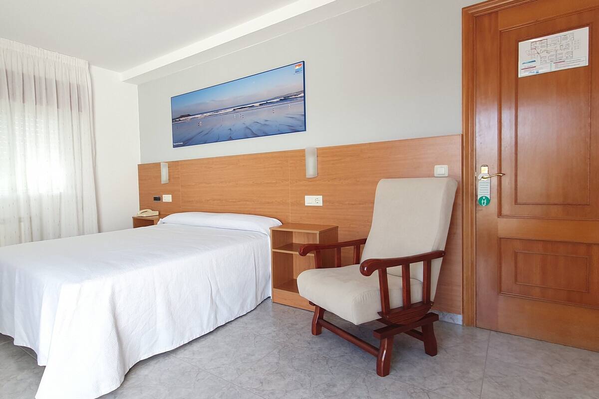 Habitación de hotel con una cama, una mesita de noche de madera, una silla, una puerta de madera y un cuadro en la pared.