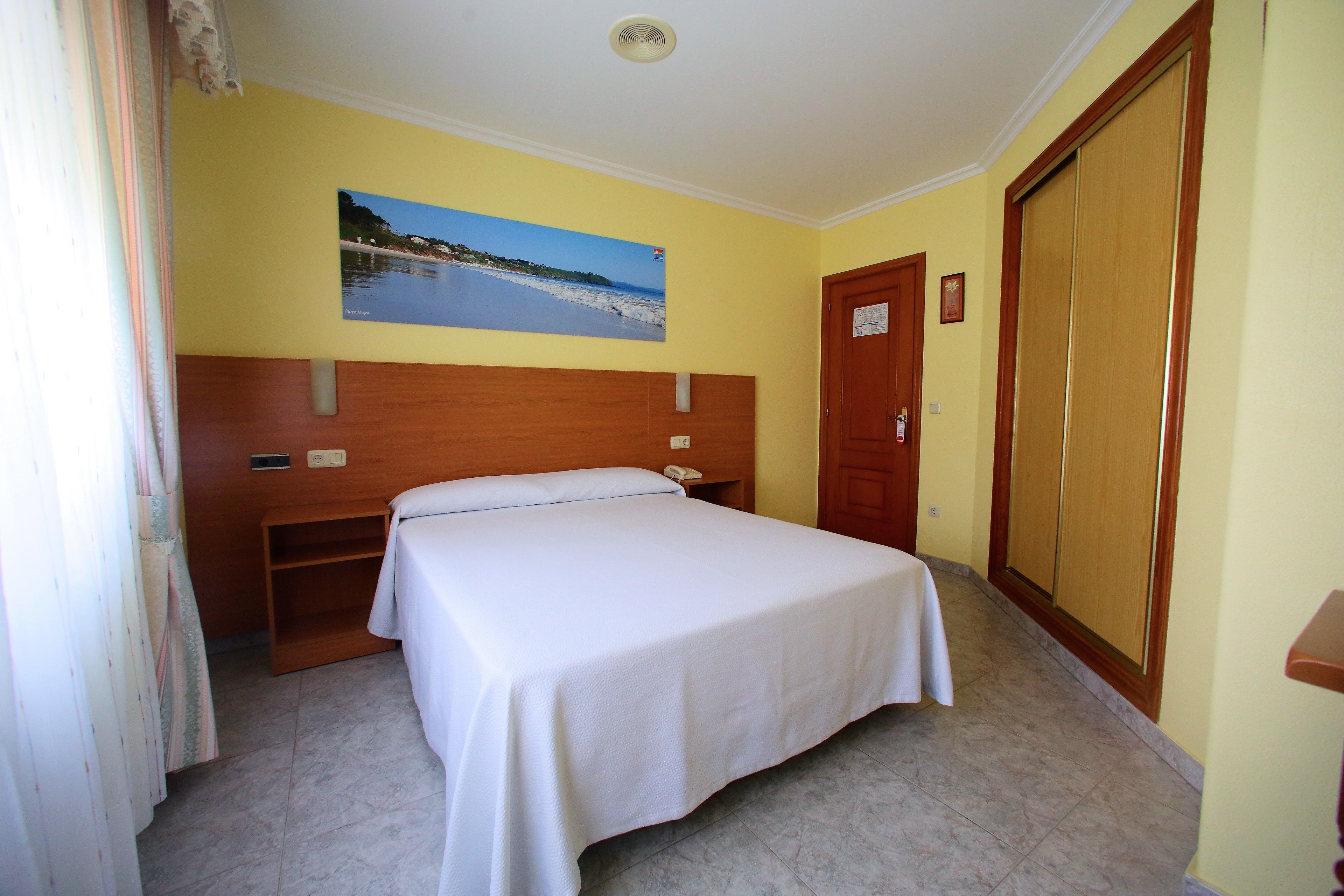 Habitación de hotel con cama, cabecera de madera, pintura de una playa, armario y escritorio con teléfono.