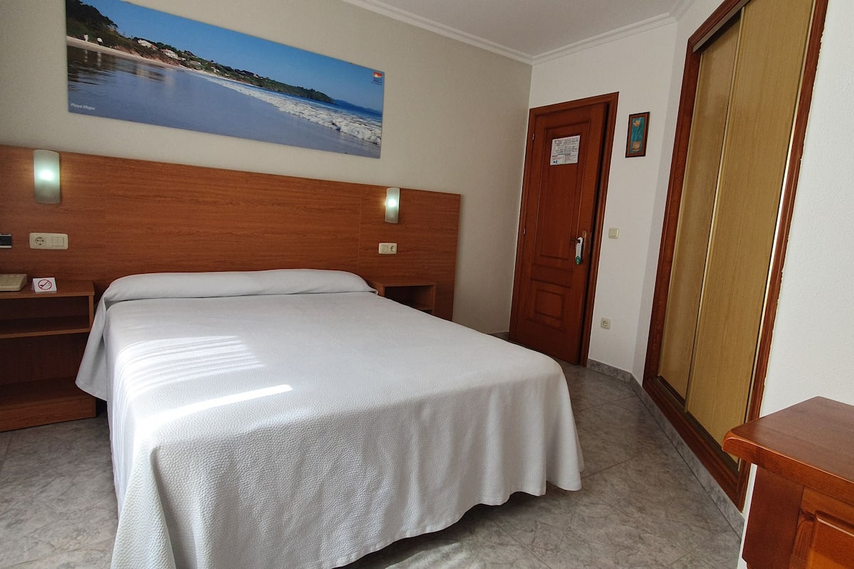 Habitación de hotel con cama, mesita de noche, teléfono, cabecera de madera, un cuadro con paisaje de playa, puerta y ventana.