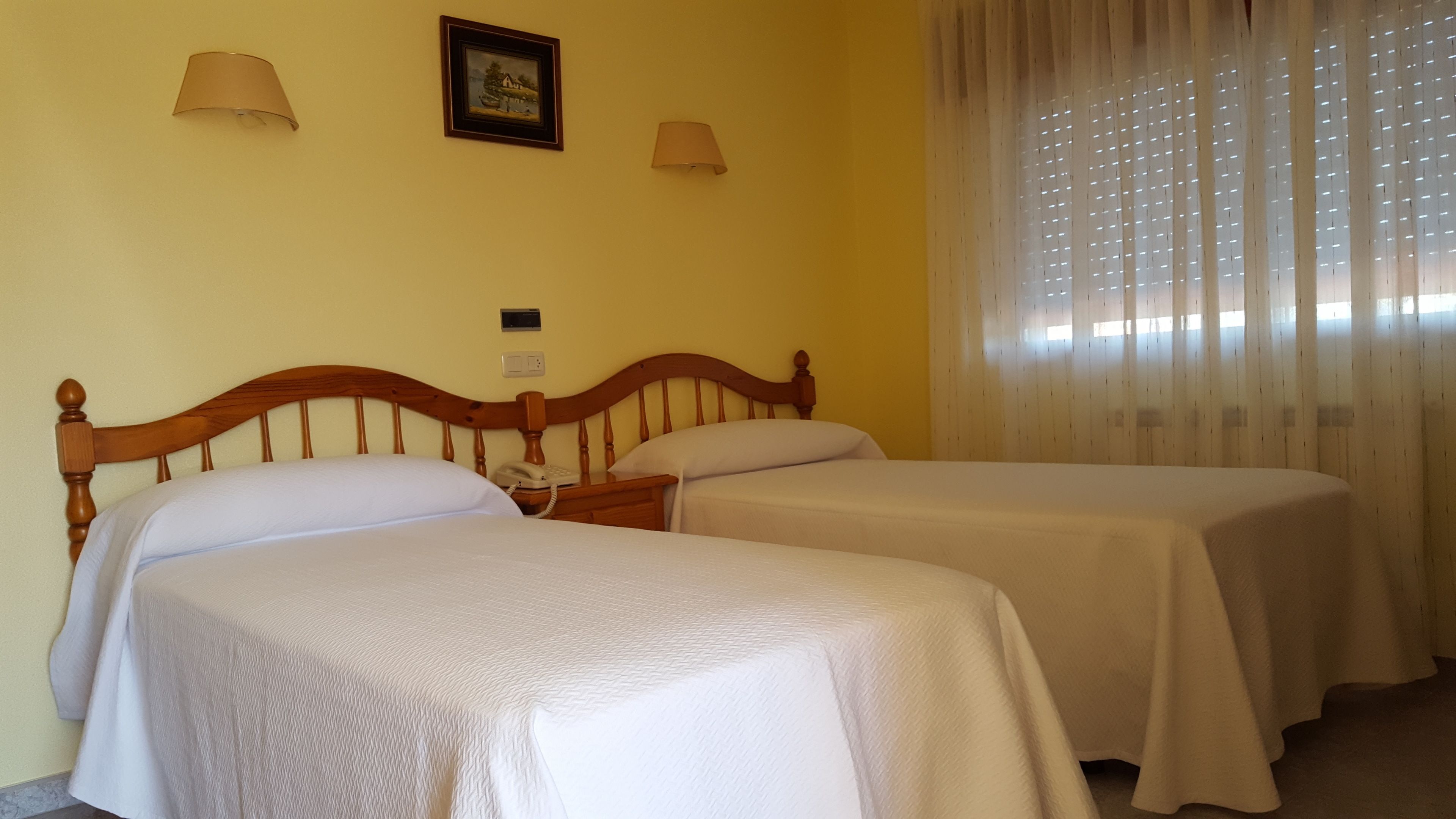 A hotel room with two single beds, a wooden headboard, a bedside telephone, and a window with sheer curtains.